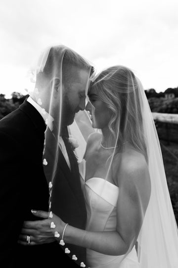 A bride and groom facing each other with their faces partially covered by a white wedding veil featuring satin heart trim. The background includes a blurred outdoor setting.
